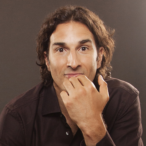 professional headshot of gary gulman that has a brown background. The man is wearing a brown shirt has shoulder length brown hair and is staring at the camera with his hand on his chin.